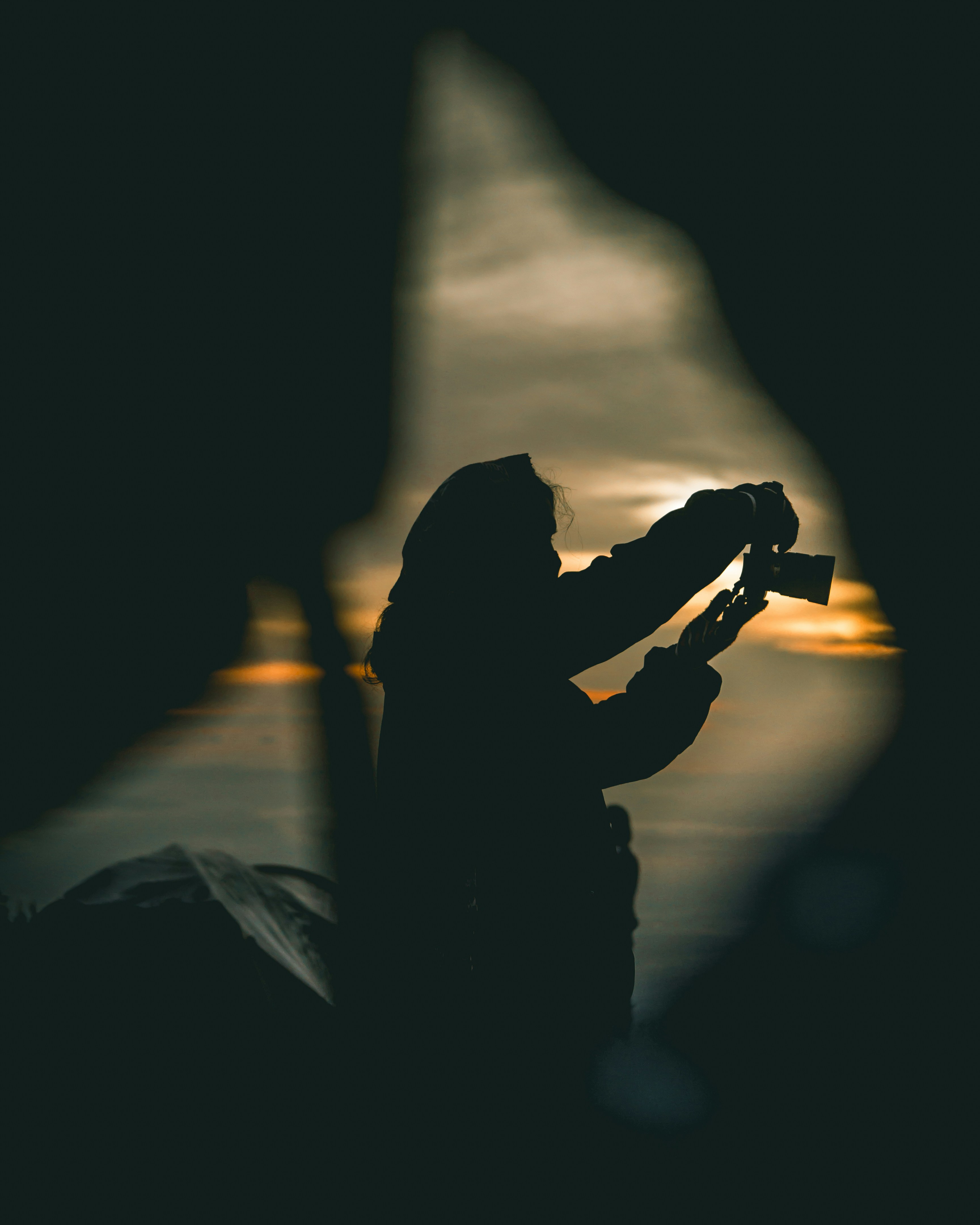 A silhouetted person adjusting a camera in amber evening light, framed by blurred dark foreground shapes.